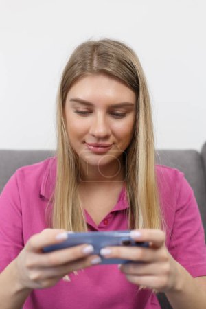 Téléchargez les photos : Portrait de gamer girl jouer jeu mobile en ligne sur téléphone intelligent. Belle femme blonde utilisant un gadget moderne pour les jeux - en image libre de droit