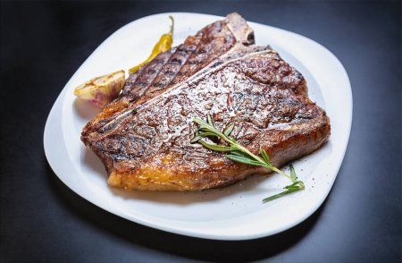 Photo for Delicious t-bone steak meat cooked on grill for dinner in American restaurant. Gourmet beefsteak grilled and served on white plate with rosemary herb - Royalty Free Image