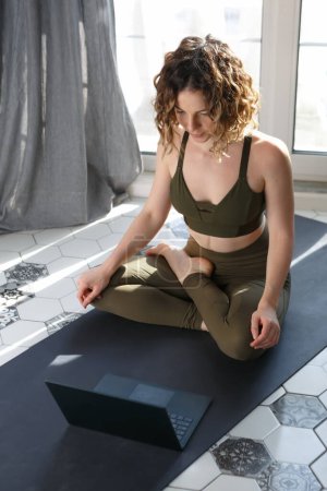 Téléchargez les photos : Young girl doing online yoga exercises at home. Beautiful white woman sitting on lotus pose in front of laptop computer - en image libre de droit