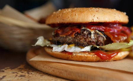 Téléchargez les photos : Grand burger américain gras servi sur une assiette en bois dans un restaurant de restauration rapide. Grand hamburger avec viande hachée cuite sur le gril et servi avec sauce ketchup et feuilles de salade de laitue dans des petits pains croustillants - en image libre de droit