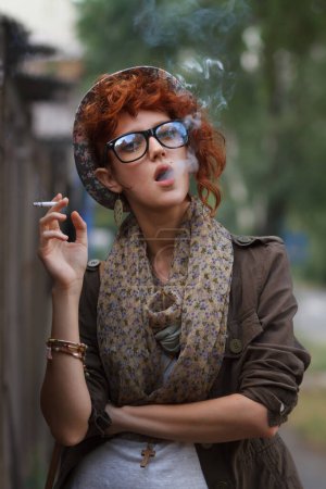 Téléchargez les photos : Teenager girl smoking a cigarette. Stylish young woman wearing fashionable retro clothes and classic glasses smokes tobacco outdoors - en image libre de droit