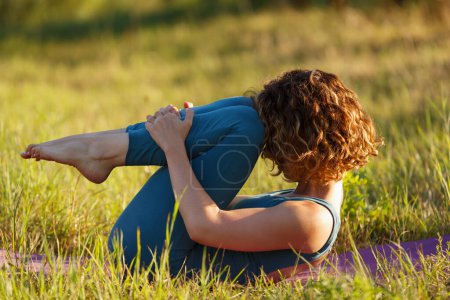 Foto de Hermosa joven haciendo yoga al aire libre al atardecer. Mujer yogini blanca con el pelo rizado estirándose en una estera en el parque - Imagen libre de derechos