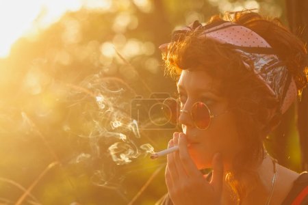 Téléchargez les photos : Cool jeune fille hipster fumant une cigarette en plein air au coucher du soleil. Portrait de modèle ukrainien élégant avec une cig dans la bouche - en image libre de droit