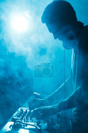 Photo for Silhoutte of club dj playing techno music set in smoke and blue lights - Royalty Free Image
