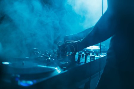 Club DJ mixing vinyl records in blue stage lights and thick smoke. Disc jockey playing music in nightclub