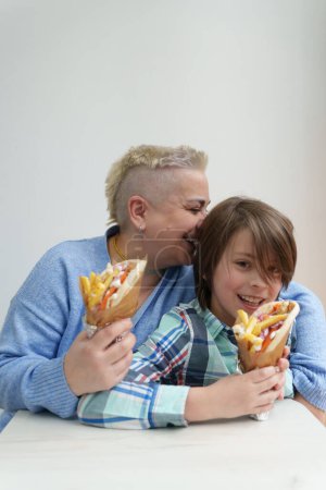 Photo for Happy mother cuddling with her son while eating gyros in Greek restaurant. Cheerful white woman with short hair and little boy enjoying their meal in a fast food diner - Royalty Free Image