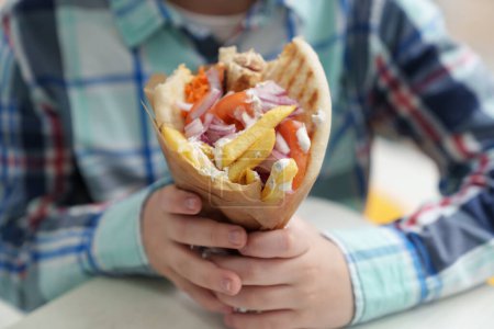 Foto de Un niño comiendo comida griega rápida en un restaurante. Foto de cerca del niño varón sosteniendo grandes giroscopios con pan de pita, carne de pollo a la parrilla, salsa tzatziki blanca, papas fritas y verduras - Imagen libre de derechos