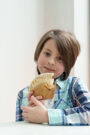 Foto de Adorable niño de edad elemental comiendo pita souvlaki con carne a la parrilla en la cafetería griega. Lindo niño blanco disfrutando de comida rápida para el almuerzo - Imagen libre de derechos