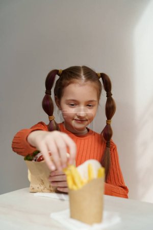 Téléchargez les photos : Joyeux blanc fille manger des frites et sandwich dans un restaurant. Portrait d'un enfant de l'âge élémentaire qui mange de la restauration rapide pour le déjeuner dans un café grec - en image libre de droit