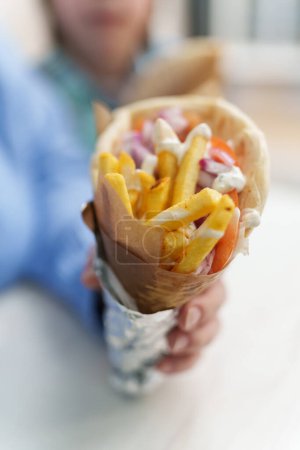 Photo for Traditional Greek gyros with pita bread, French fries, tzatziki sauce, grilled chicken meat and fresh vegetables. Unrecognizable female person holds big wrapped pita souvlaki in hand - Royalty Free Image