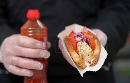 Foto de El cocinero sosteniendo perro caliente exótico en la mano. Chef cocinó sándwich de perritos calientes con carne de pollo marinada cocinada a la parrilla y servida con verduras, hierbas de orégano y salsa asiática picante - Imagen libre de derechos
