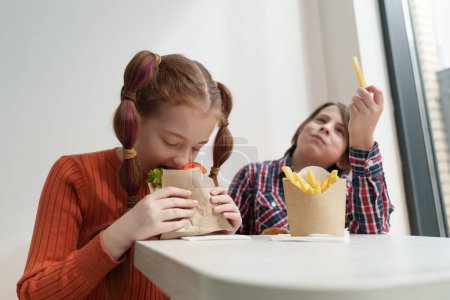 Téléchargez les photos : Deux mignons enfants d'âge élémentaire qui mangent de la restauration rapide pour le déjeuner. Deux petits enfants ukrainiens dégustant le repas dans un café européen - en image libre de droit
