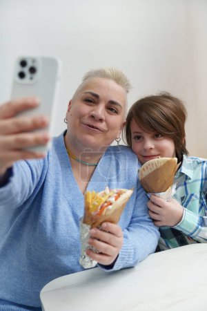 Foto de Feliz familia ucraniana disfrutando de la comida en un café europeo. Mujer blanca adulta tomando una selfie junto con su hijo mientras sostiene la merienda griega gyros en las manos - Imagen libre de derechos
