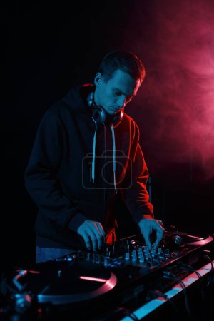 Téléchargez les photos : Jeune DJ cool jouant des vinyles sur la fête. Disk jokey mélangeant des pistes musicales dans une boîte de nuit sombre - en image libre de droit