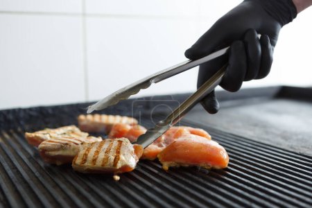 Foto de Cocinar con guantes negros convierte la carne en parrilla con pinzas. Chef asando filete de pollo en una sartén caliente en una cocina de restaurante - Imagen libre de derechos