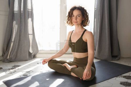 Photo for Beautiful yogi girl sitting in lotus pose on the mat. Attractive white woman with curly hair meditating at home after yoga workout - Royalty Free Image