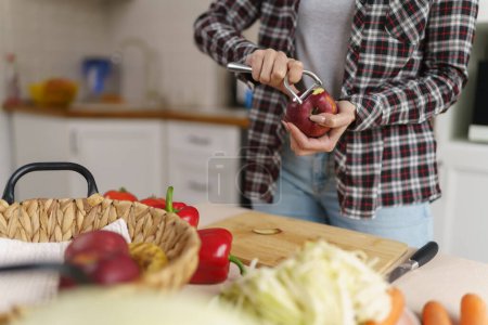 Téléchargez les photos : Femme épluchant une pomme rouge avec un éplucheur. Femme cuisinant le déjeuner dans une cuisine domestique - en image libre de droit