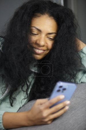 Téléchargez les photos : Heureuse femme noire communiquant en ligne avec l'application de téléphone mobile. Portrait de la personne à la peau foncée gaie en utilisant l'application smartphone - en image libre de droit