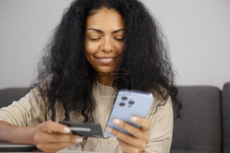 Téléchargez les photos : Heureuse femme noire payant par carte de crédit en ligne. Enthousiaste personne à la peau foncée faisant du shopping dans l'application mobile sur smartphone - en image libre de droit