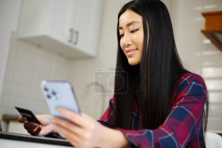 Photo for Happy Asian girl buying online with credit card and mobile app. Cheerful Vietnamese female person shopping in internet store with smartphone - Royalty Free Image