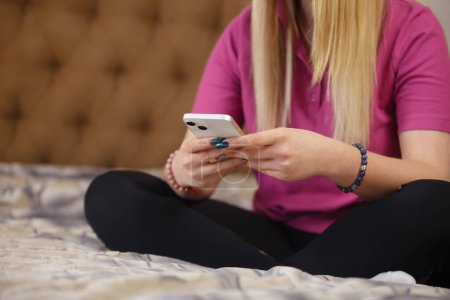 Foto de Mujer joven escribiendo mensaje en el teléfono móvil. Mujer blanca sentada en la cama y la navegación aplicación de teléfono inteligente moderno - Imagen libre de derechos