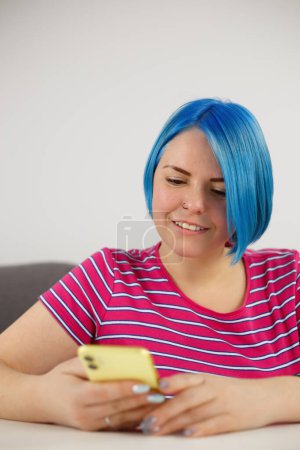 Foto de Feliz mujer blanca escribiendo mensaje en el teléfono inteligente amarillo. Alegre azul de pelo femenino de navegación aplicación de teléfono móvil - Imagen libre de derechos