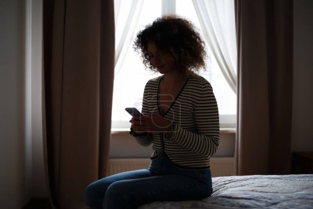 Foto de Mujer joven escribiendo mensaje en el teléfono móvil en el cuarto oscuro. Blanco persona femenina mensajes de texto en línea con teléfono inteligente desde el dormitorio - Imagen libre de derechos