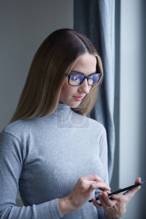 Foto de Hermosa mujer rubia en camisa de cuello alto gris navegador tableta. Joven mujer blanca un gafas usando gadget portátil con internet inalámbrico - Imagen libre de derechos
