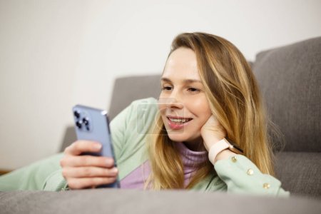 Happy white woman browsing smartphone app while lying on couch. Beautiful adult female dating online with mobile application. Cheerful Ukrainian person communicating online with cellphone
