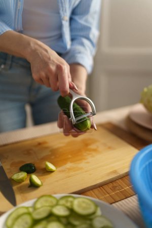 Photo for Housewife peeling off cucumber with a peeler tool in close up. Female person peels vegetables for lunch at home - Royalty Free Image