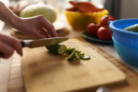 Photo for Female hand chopping cucumber with a sharp knife on a wooden cutting board in close up. Woman cooking healthy lunch meal at home with fresh vegetables - Royalty Free Image