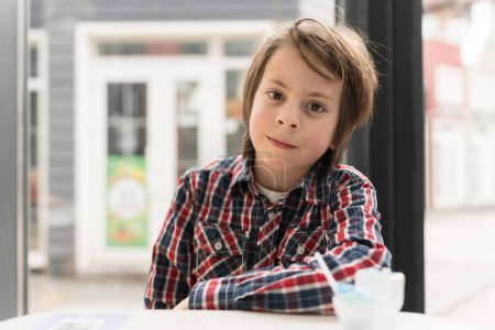 Foto de Retrato de un lindo chico blanco con una camisa a cuadros posando en un café. Niño hambriento esperando orden en un restaurante de comida rápida - Imagen libre de derechos
