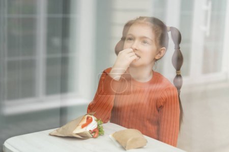 Photo for Little white girl with ponytails eating fast food in a cafe. Beautiful child eats fries and sandwich in a diner, photographed through a window - Royalty Free Image
