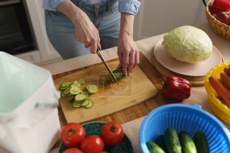 Téléchargez les photos : Des femmes coupent des concombres avec un couteau sur une table de cuisine. Femme cuisine sain déjeuner végétarien et peelings de compostage dans un bac bokashi - en image libre de droit