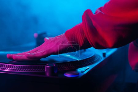 Téléchargez les photos : Hand of a hip hop DJ scratching vinyl record on turntable in close up. Club disc jockey mixant des disques sur la fête dans une épaisse fumée et des lumières de scène - en image libre de droit