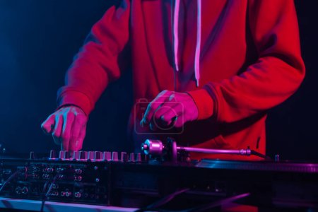 Foto de Club DJ ajustando el volumen en un dispositivo mezclador de sonido en el club nocturno. Foto de cerca del disc jockey profesional con capucha roja tocando música en una fiesta - Imagen libre de derechos