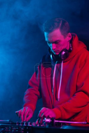 Photo for Cool young hip hop DJ in red hoodie mixing vinyl records on a party in night club. Professional disc jockey plays set in smoke on stage - Royalty Free Image