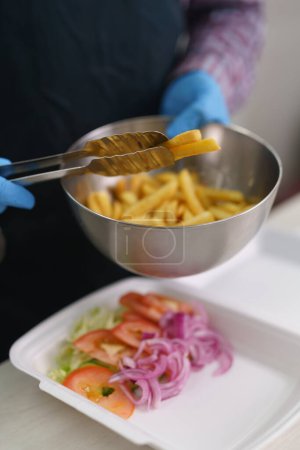 Téléchargez les photos : Cook a mis des frites dans une boîte à lunch. Cuisinier préparant un plat à emporter dans un restaurant grec de restauration rapide - en image libre de droit