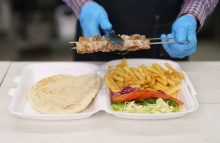 Téléchargez les photos : Cuisiner en préparant un plat traditionnel grec kalamaki dans une cuisine de restaurant. Chef mettre la viande de porc grillée avec pinces dans une boîte à lunch avec du pain pita, des frites et des légumes frais - en image libre de droit