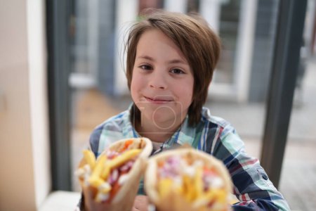Téléchargez les photos : Joyeux petit garçon dans un fast-food. Portrait d'enfant blanc joyeux prêt à déjeuner dans un restaurant grec - en image libre de droit