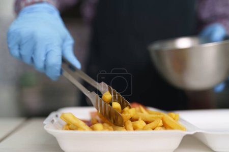 Téléchargez les photos : Le cuisinier sert des frites avec des pinces métalliques dans une boîte à lunch. Chef préparant la restauration rapide à emporter dans un restaurant grec - en image libre de droit