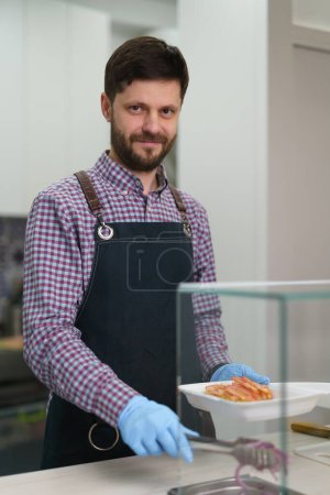 Foto de Hombre blanco guapo trabajando en un restaurante griego de comida rápida. Retrato de un hombre alegre sirviendo lonchera para llevar en un restaurante - Imagen libre de derechos