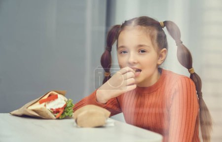 Photo for Beautiful white girl with ponytails eating fast food. Portrait of a cute elementary age child enjoying fries for lunch, photographer in a restaurant through a window - Royalty Free Image