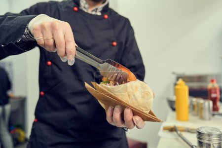 Foto de El cocinero que prepara el plato griego pita souvlaki en un restaurante mediterráneo. Chef puso tomate en rodajas en un sándwich - Imagen libre de derechos