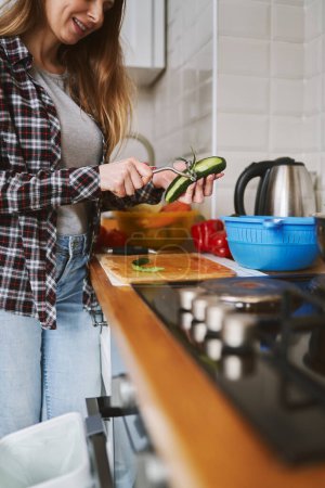 Photo for Cheerful adult woman peeling off a cucumber skin with a peeler tool. Female person cooking fresh salad at home - Royalty Free Image