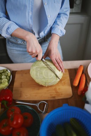 Photo for Woman cutting a cabbage with a knife on a wooden board. Female person preparing healthy vegetarian salad with vegetables - Royalty Free Image