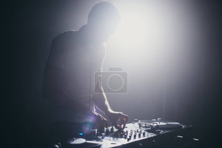 Foto de Silueta de un DJ del club mezclando discos de vinilo en el escenario. Escena retroiluminada con un disc jockey tocando música en un concierto en un club nocturno - Imagen libre de derechos