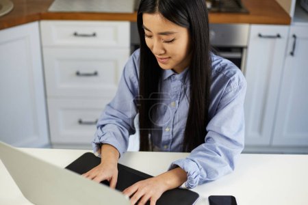 Photo for Cheerful young Asian student studying online. Beautiful POC female from Vietnam typing text on a modern laptop computer at home - Royalty Free Image