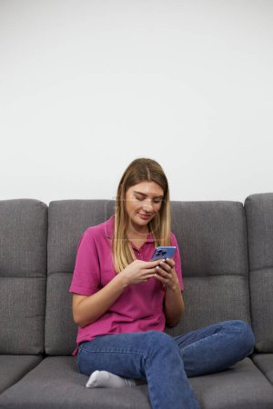 Photo for Pretty blonde female sitting on a couch at home and typing a message on a smart phone with a smile - Royalty Free Image