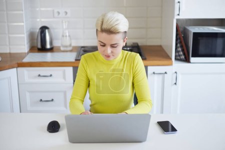 Photo for Young woman working on a computer at home. Freelancer female person typing text on a laptop in a domestic kitchen. - Royalty Free Image